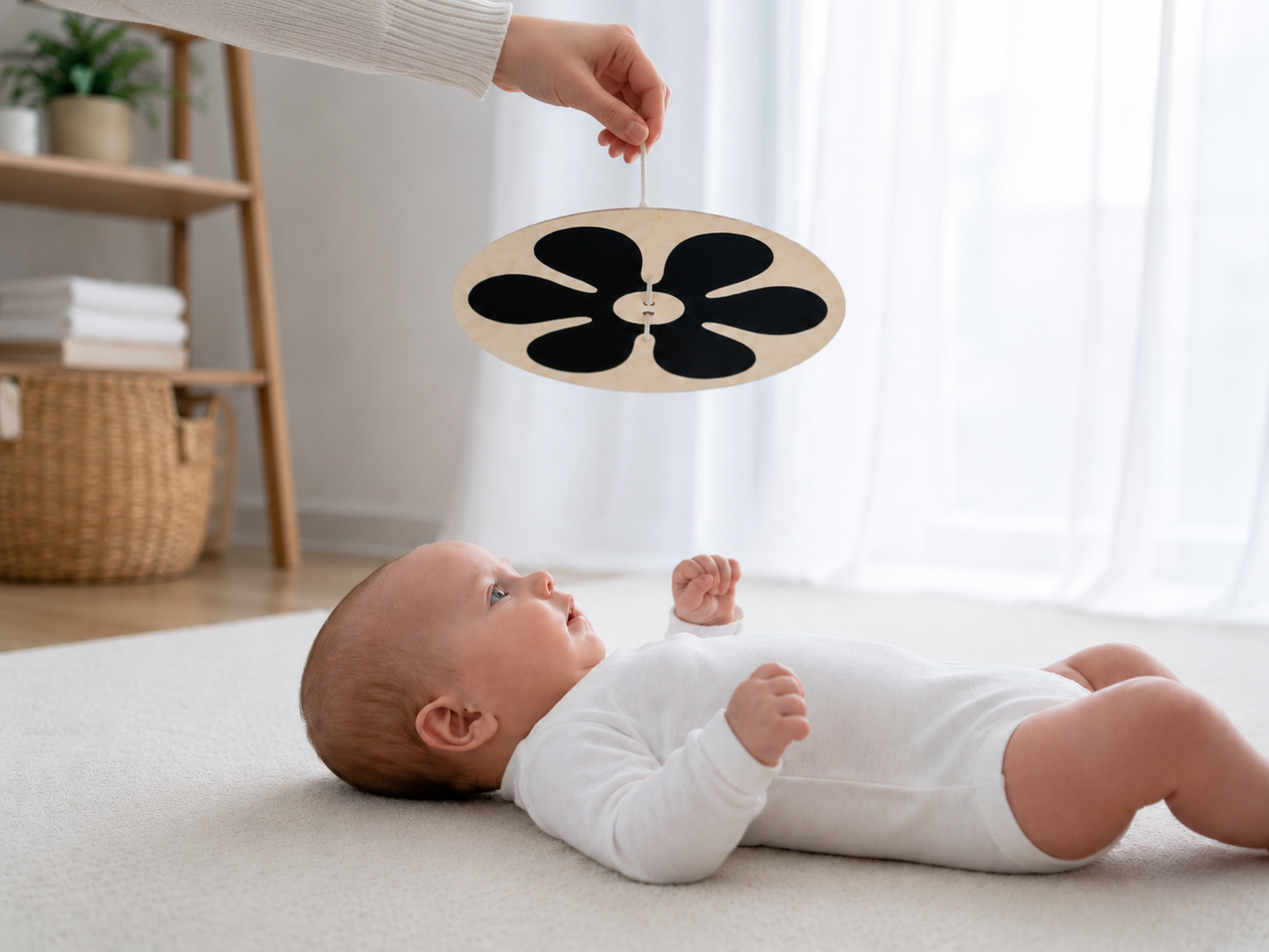 A baby in a white onesie lies on a cream rug, reaching toward a black flower-shaped baby toy hanging from an adult's hand in a bright, modern nursery.