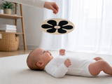 A baby in a white onesie lies on a cream rug, reaching toward a black flower-shaped baby toy hanging from an adult's hand in a bright, modern nursery.